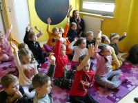 Grupa dzieci wraz z opiekunem siedzą na dywanie i podnoszą rękę, a by udzielić odpowiedzi na pytanie prowadzącej zajęcia