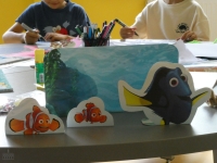 Na pierwszym planie ustawiono kartonowe figurki rybek, w tle widać dwoje dzieci malujących flamastrami
