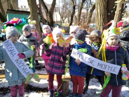 Grupa przedszkolaków stojąca na podwórku wśród drzew trzyma w dłoniach kolorowe paski bibuły i napisy „Wiosna, wiosna”, „Urodziły się motyle”
