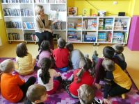 Grupa dzieci w wieku przedszkolnym siedzi na dywanie i słucha opowiadania, które czyta bibliotekarka