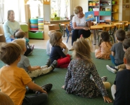 Grupa dzieci siedzi na dywanie i słucha opowiadania czytanego przez bibliotekarkę