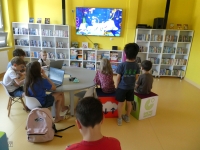 Grupa dzieci uczestniczy w wakacyjnych zajęciach, grając na konsoli Nintendo Switch, tabletach i laptopach