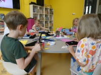 Dzieci siedzą przy stoliku, wykonując prace plastyczne z wykorzystaniem książek, materiału, pianek i drucików kreatywnych